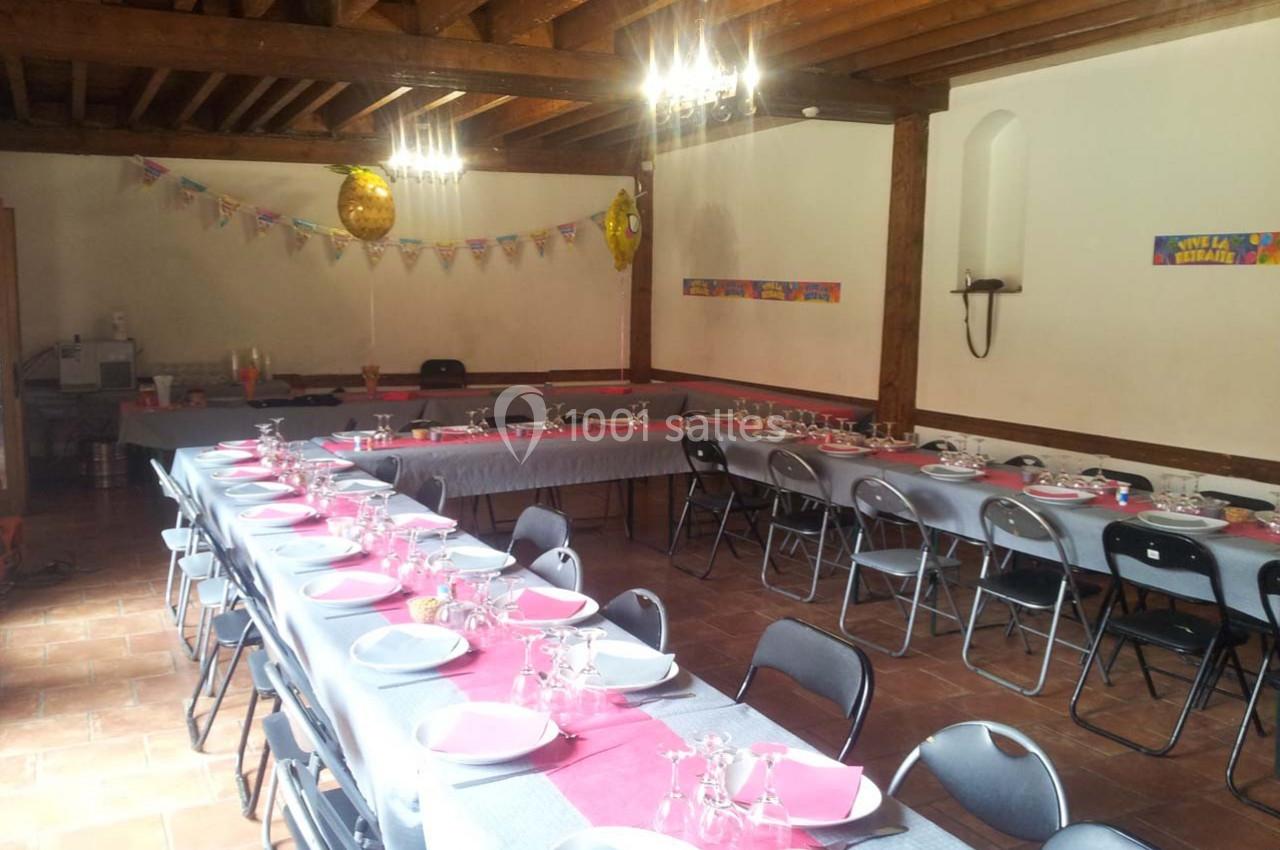 Salle décorée pour une fête avec longues tables dressées, nappes grises, assiettes roses et guirlandes colorées.