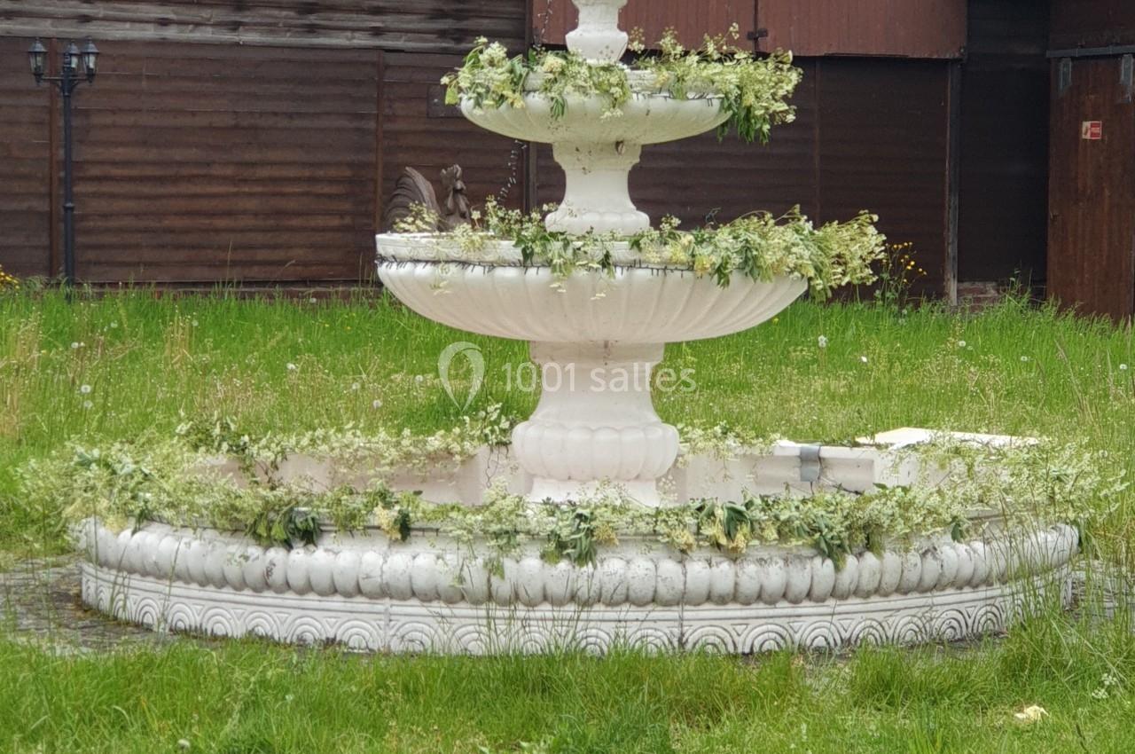 Fontaine blanche à trois niveaux ornée de végétation, avec une statue d'enfant au sommet, située sur une pelouse devant un…