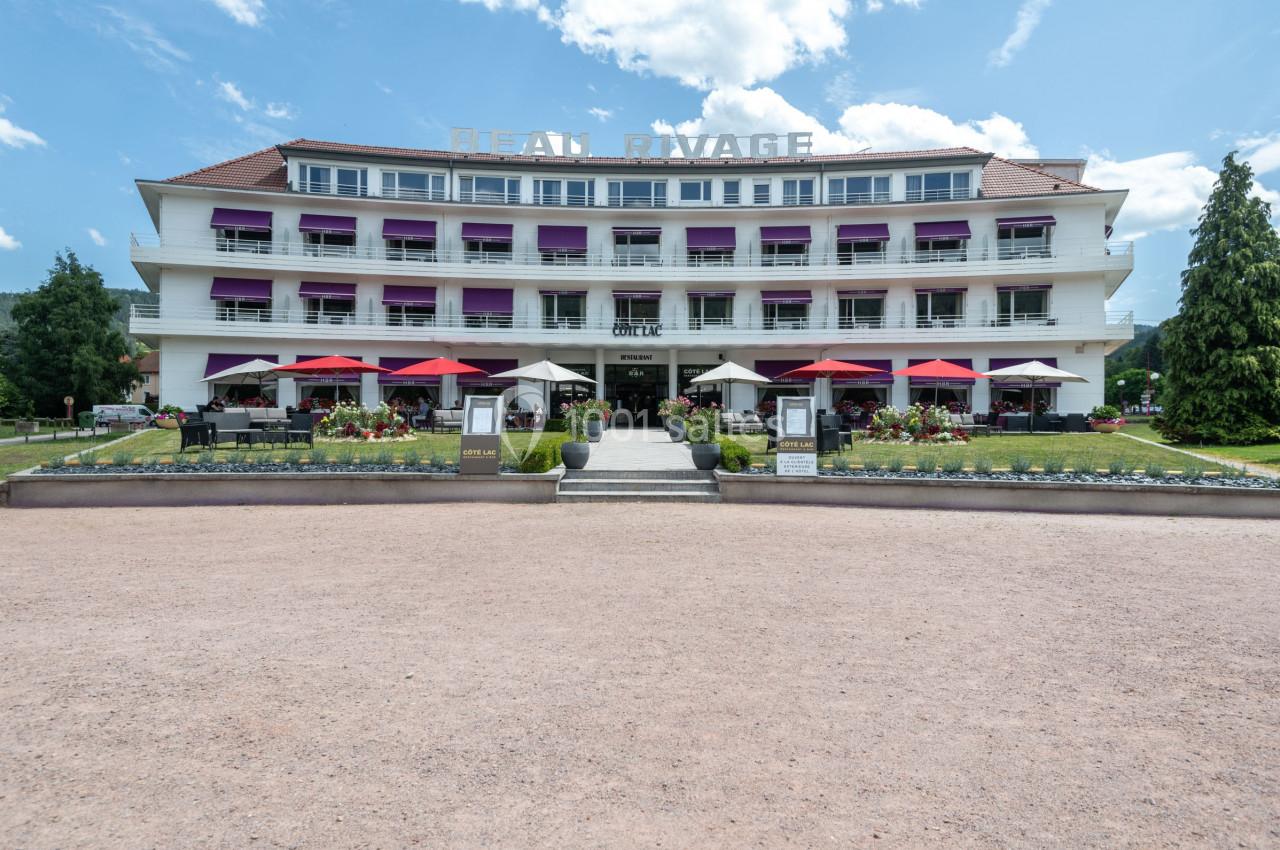 Façade d'un hôtel avec balcons, stores violets et terrasse extérieure équipée de parasols rouges.