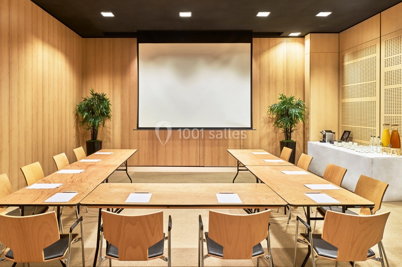 Salle de réunion avec tables en U, chaises en bois, écran de projection, plantes et buffet avec boissons.