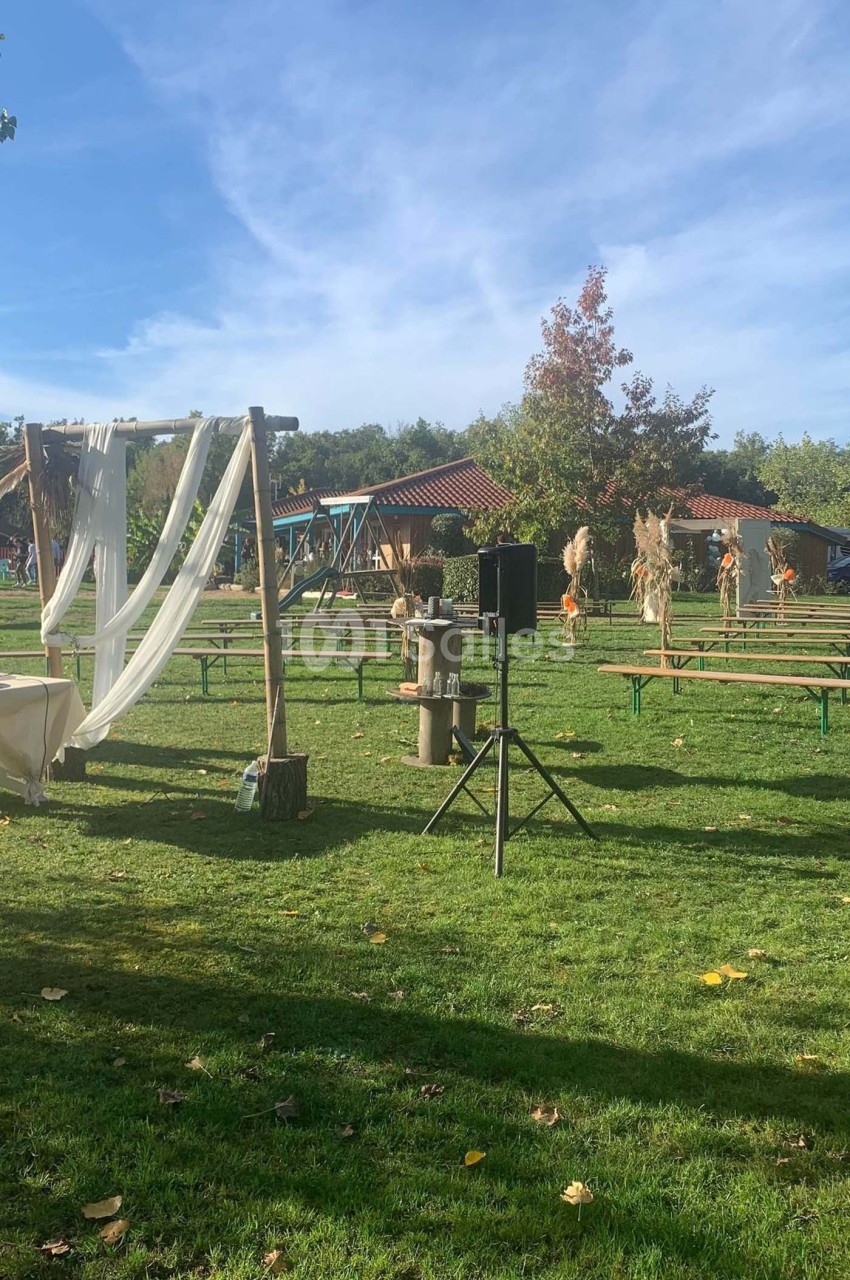 Espace extérieur avec bancs en bois, arche décorée de voilages blancs et pelouse sous un ciel dégagé.