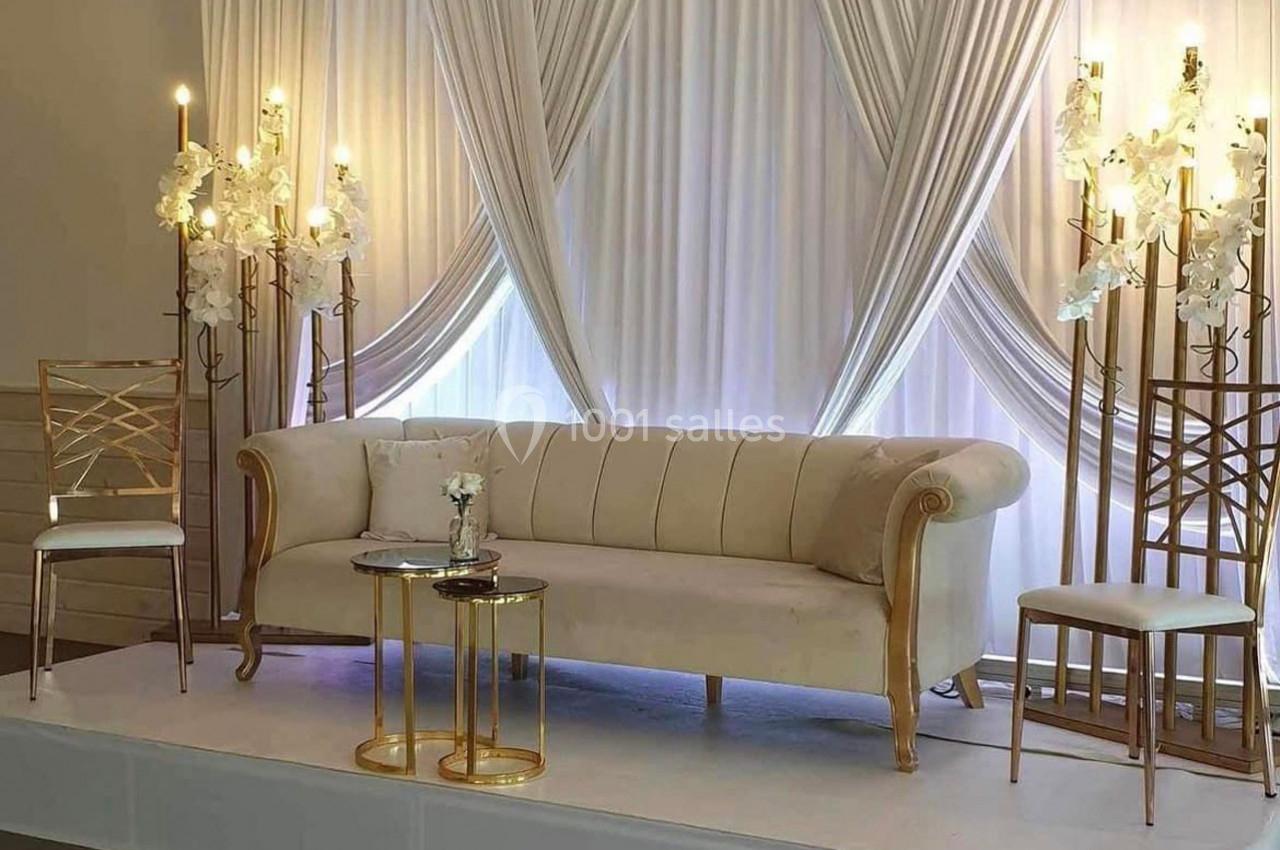 Canapé beige avec accoudoirs dorés, flanqué de chaises et de chandeliers ornés de fleurs blanches, devant des rideaux…
