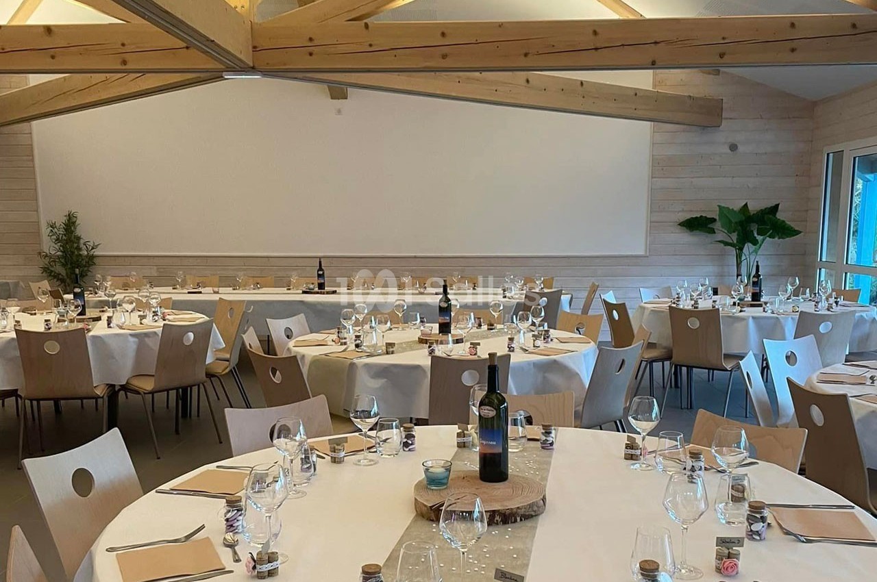 Salle de réception lumineuse avec tables rondes dressées, nappes blanches, chaises en bois et décoration sobre.