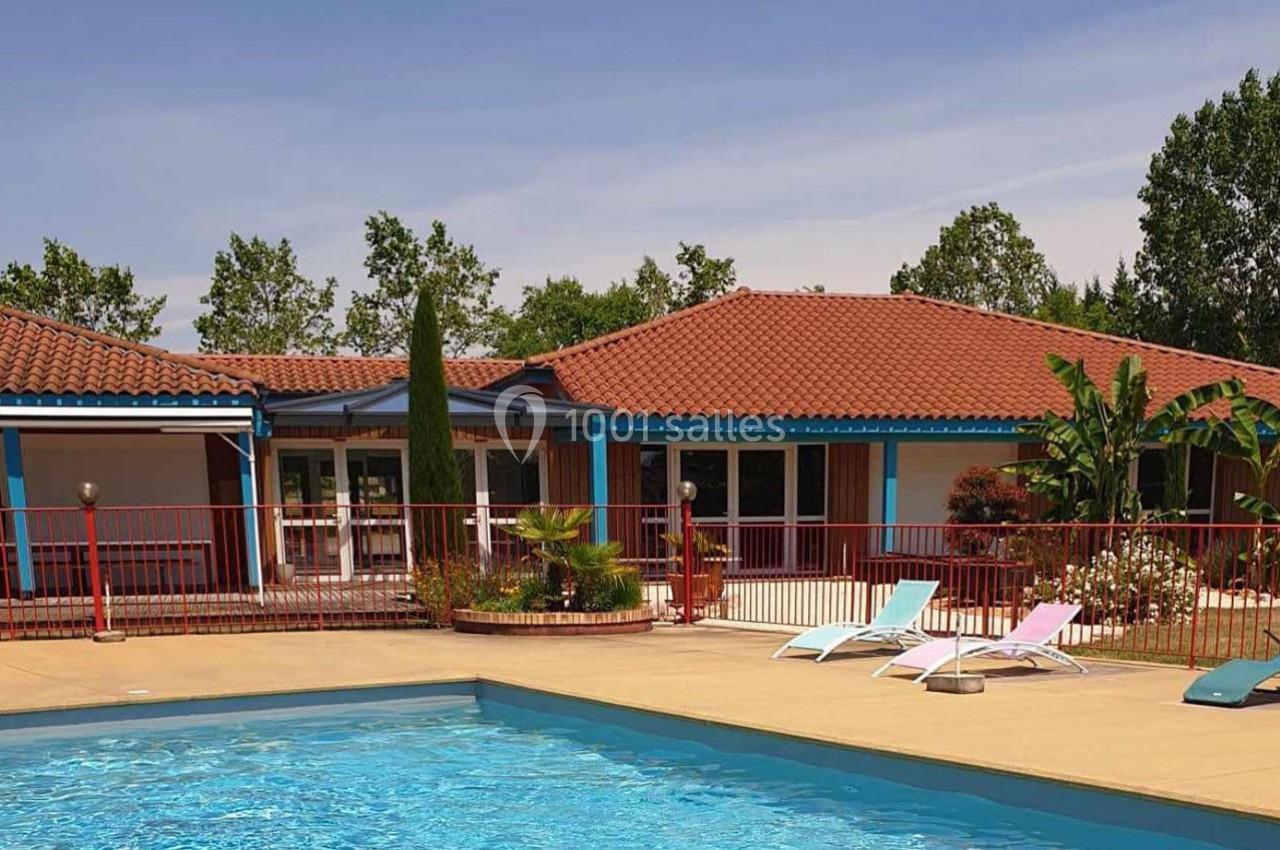 Maison avec toit en tuiles rouges, terrasse, jardin arboré et piscine entourée de chaises longues colorées.