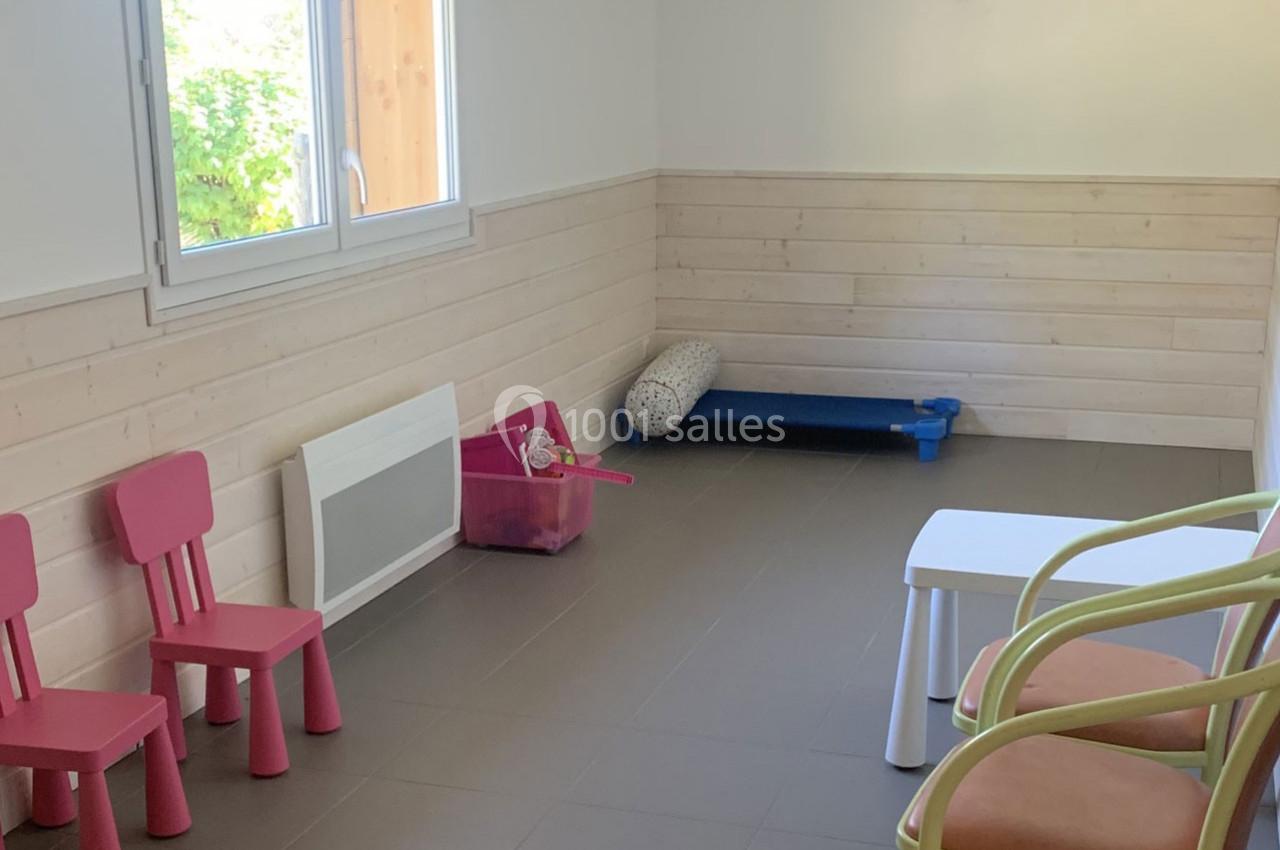 Salle lumineuse avec chaises roses pour enfants, table blanche, tapis bleu et fenêtre donnant sur un espace vert.