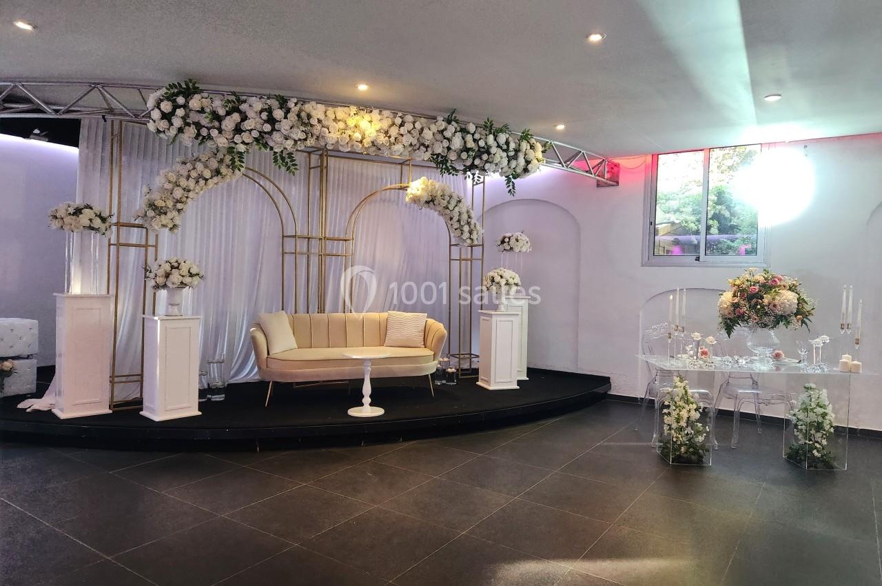 Location salle Roquevaire (Bouches-du-Rhône) - Nao Athéna #2 Décoration de salle avec arche florale blanche, canapé beige, table transparente et éclairage naturel.