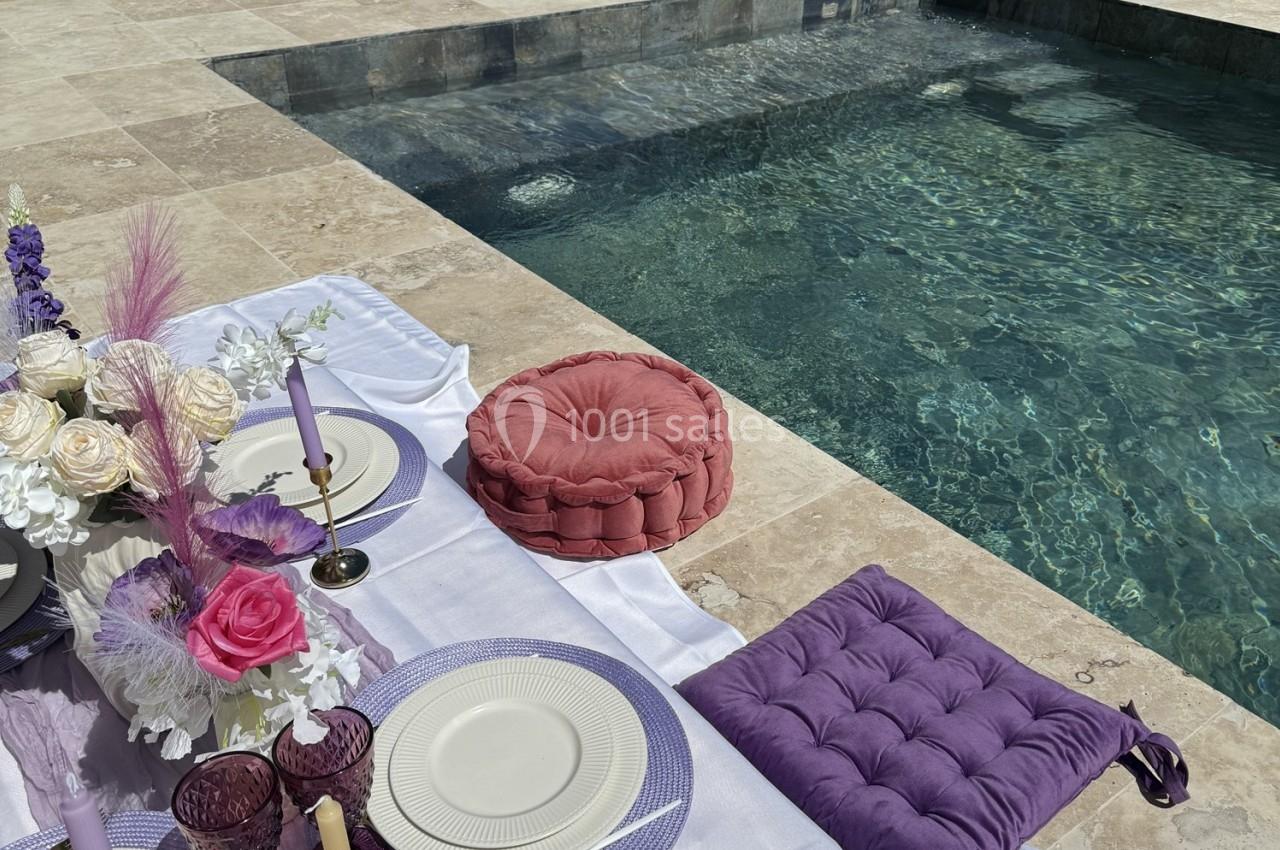 Location salle Roquevaire (Bouches-du-Rhône) - Nao Athéna #8 Table de pique-nique élégante avec coussins colorés et vaisselle raffinée, disposée au bord d'une piscine ensoleillée.