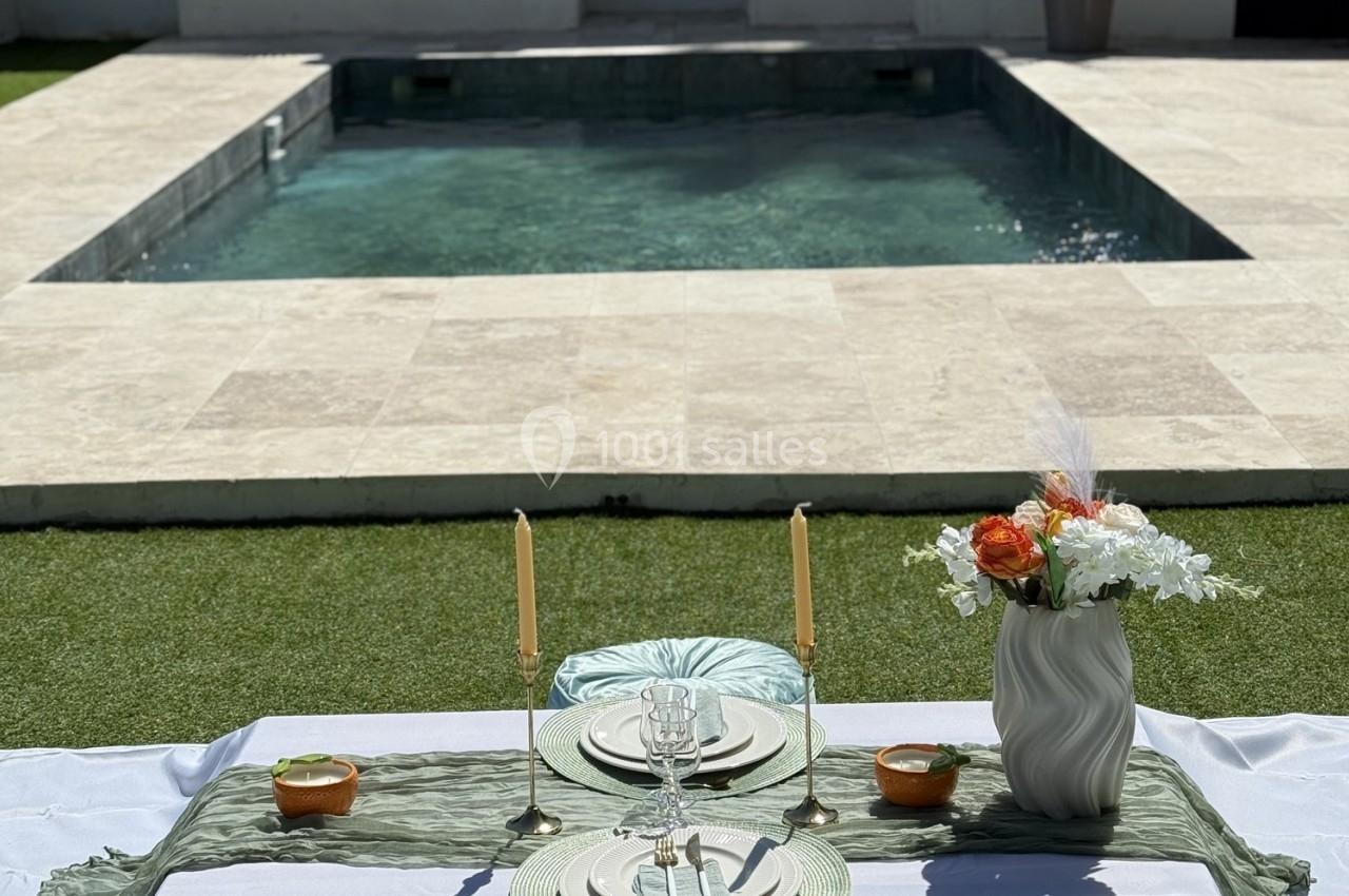 Location salle Roquevaire (Bouches-du-Rhône) - Nao Athéna #10 Table dressée avec vaisselle élégante, bougies et fleurs, disposée devant une piscine entourée de pierre et d'herbe.