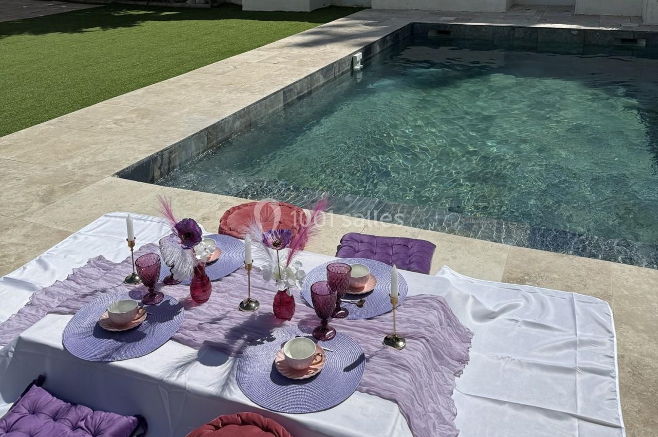 Location salle Roquevaire (Bouches-du-Rhône) - Nao Athéna #16 Table dressée avec vaisselle rose et violette, décorée de plumes et fleurs, près d'une piscine et d'une pelouse.