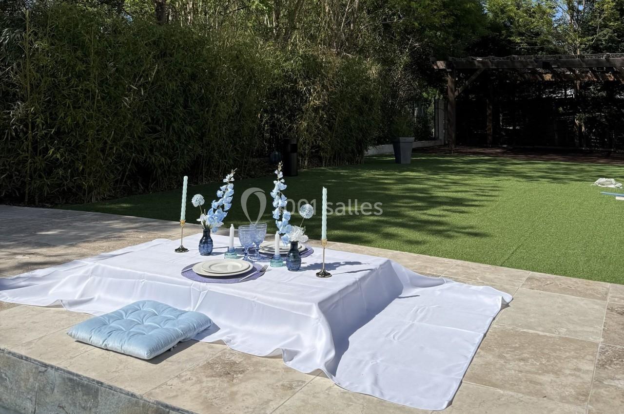 Location salle Roquevaire (Bouches-du-Rhône) - Nao Athéna #23 Table basse dressée sur une nappe blanche avec des bougies et des fleurs bleues, située en extérieur près d'une pelouse.