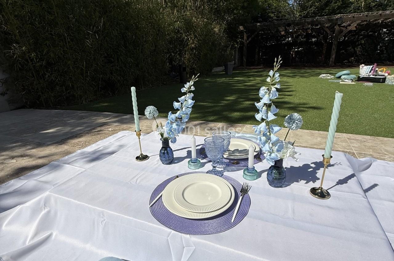 Location salle Roquevaire (Bouches-du-Rhône) - Nao Athéna #24 Table extérieure dressée avec vaisselle blanche, bougies et fleurs bleues, sur une nappe blanche dans un jardin.