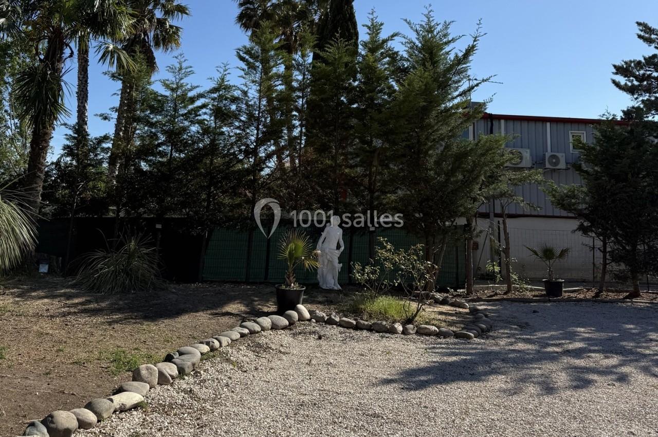 Location salle Roquevaire (Bouches-du-Rhône) - Nao Athéna #28 Allée gravillonnée bordée de pierres, avec une statue blanche entourée d'arbres et un bâtiment en arrière-plan.