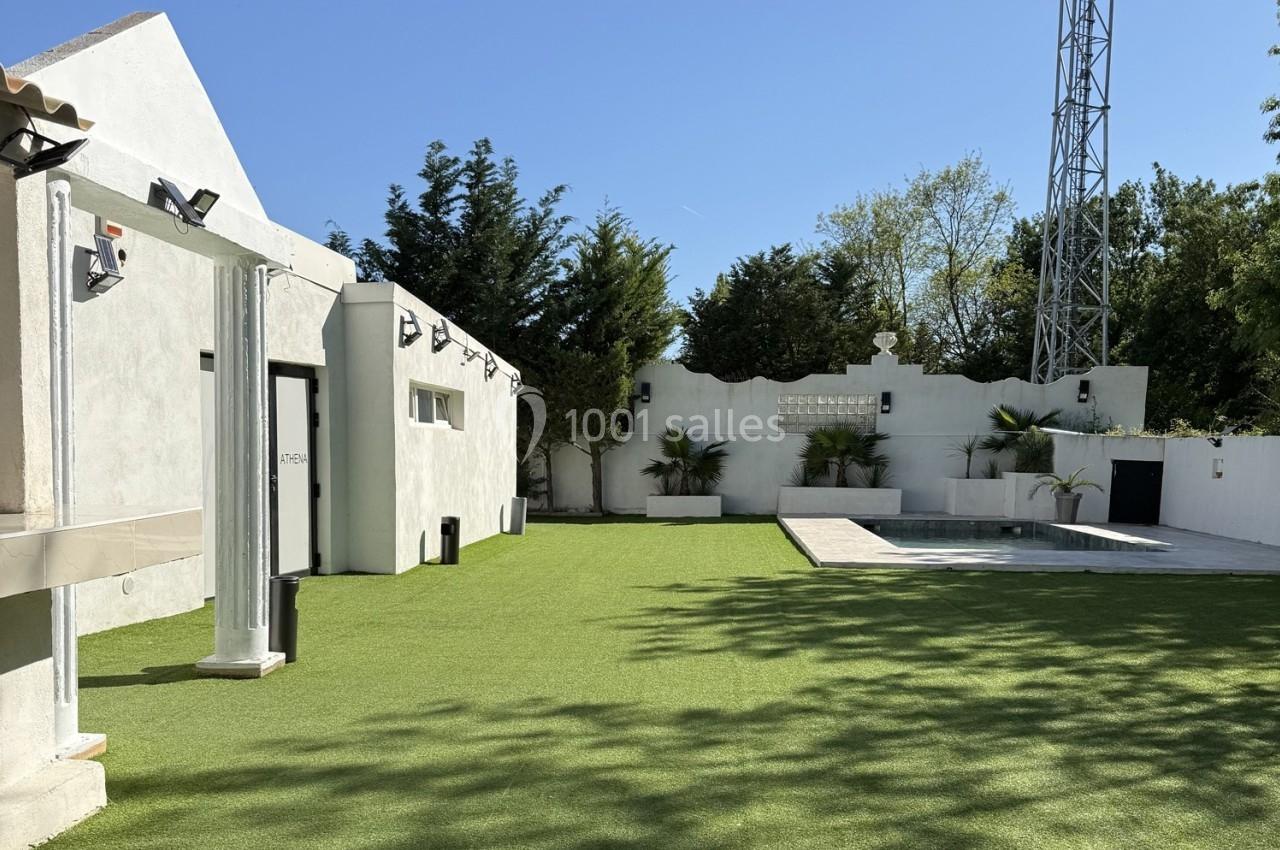Location salle Roquevaire (Bouches-du-Rhône) - Nao Athéna #32 Jardin avec pelouse synthétique, petite piscine, murs blancs et arbres en arrière-plan sous un ciel dégagé.
