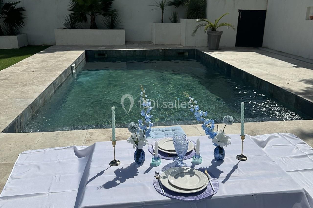Location salle Roquevaire (Bouches-du-Rhône) - Nao Athéna #37 Table dressée avec vaisselle et fleurs bleues près d'une piscine entourée de dalles claires et de végétation.