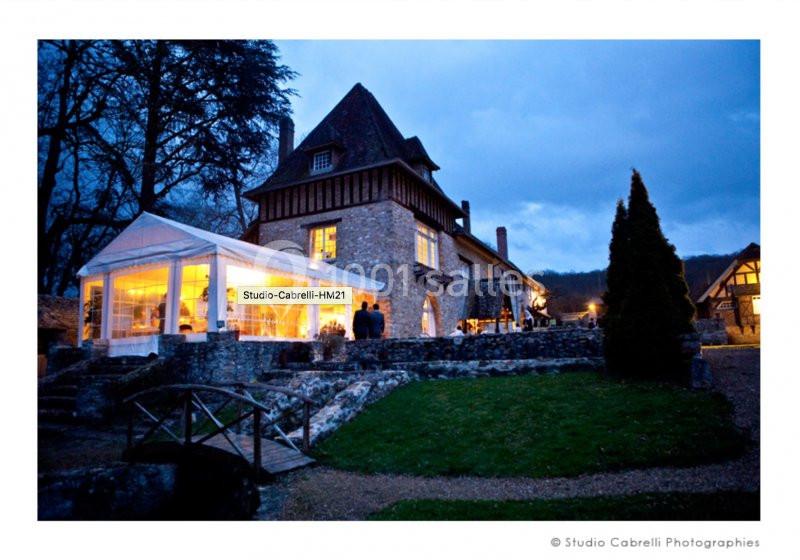 Bâtiment en pierre avec une verrière éclairée, entouré d'arbres et d'un chemin en soirée.