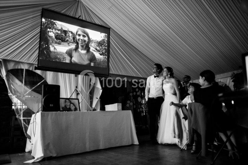 Un couple de mariés regarde une projection vidéo dans une salle décorée, entourés de quelques invités.
