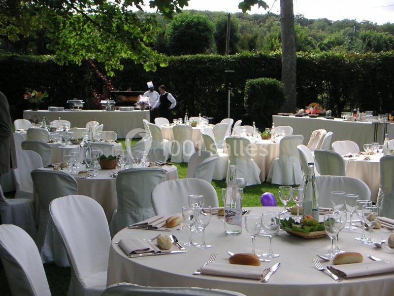 Tables rondes dressées avec nappes blanches pour un repas en extérieur, entourées de verdure et de personnel en arrière-plan.