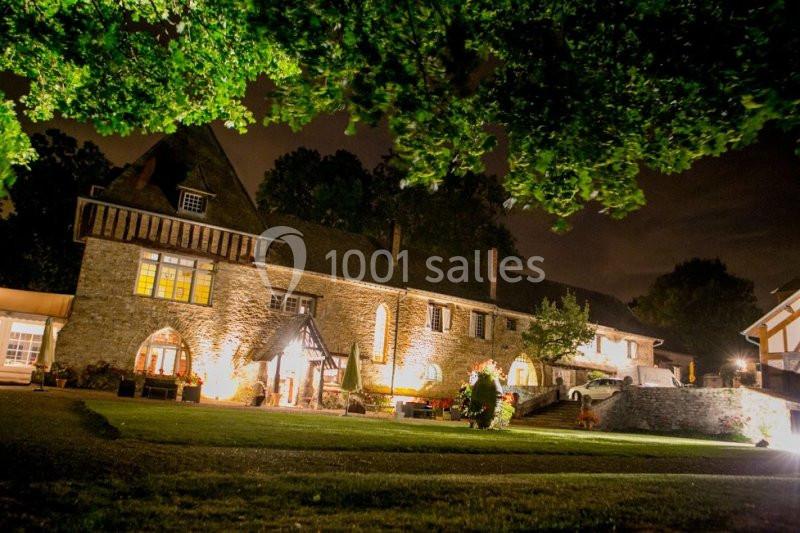 Vue nocturne d'une grande bâtisse en pierre éclairée, entourée d'arbres et d'une pelouse bien entretenue.
