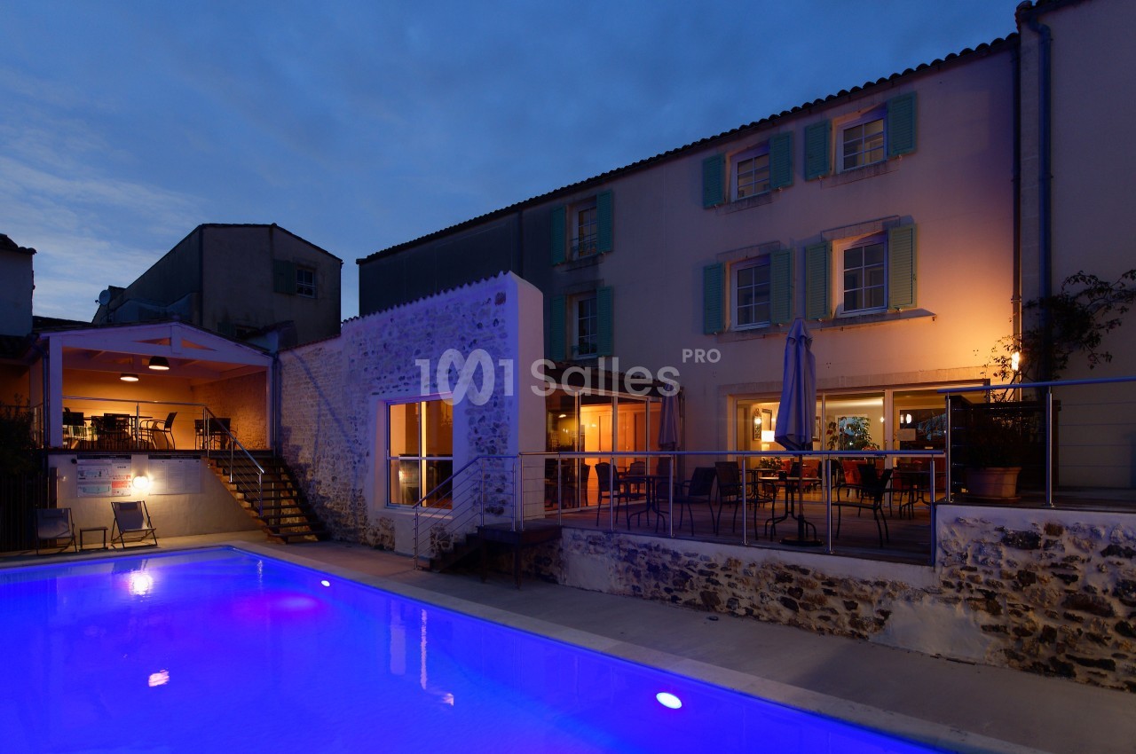Maison en pierre éclairée à la tombée de la nuit avec piscine illuminée en premier plan et terrasse aménagée.