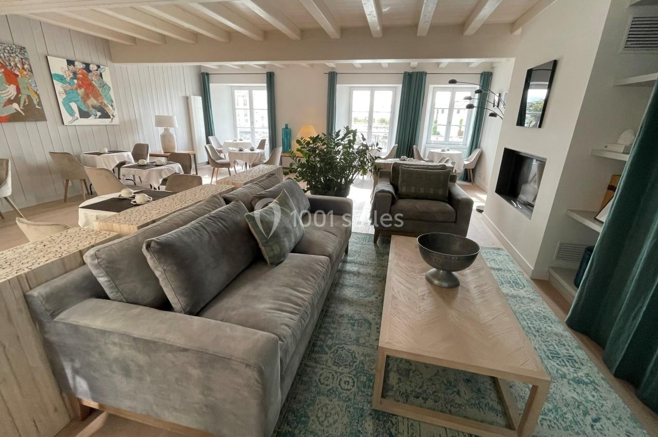 Salon lumineux avec canapé gris, table basse en bois, tapis turquoise et vue sur une salle à manger et des fenêtres.