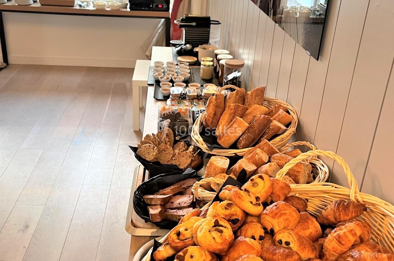 Buffet de petit-déjeuner avec viennoiseries, pains variés, confitures et boissons disposés sur une table en bois.