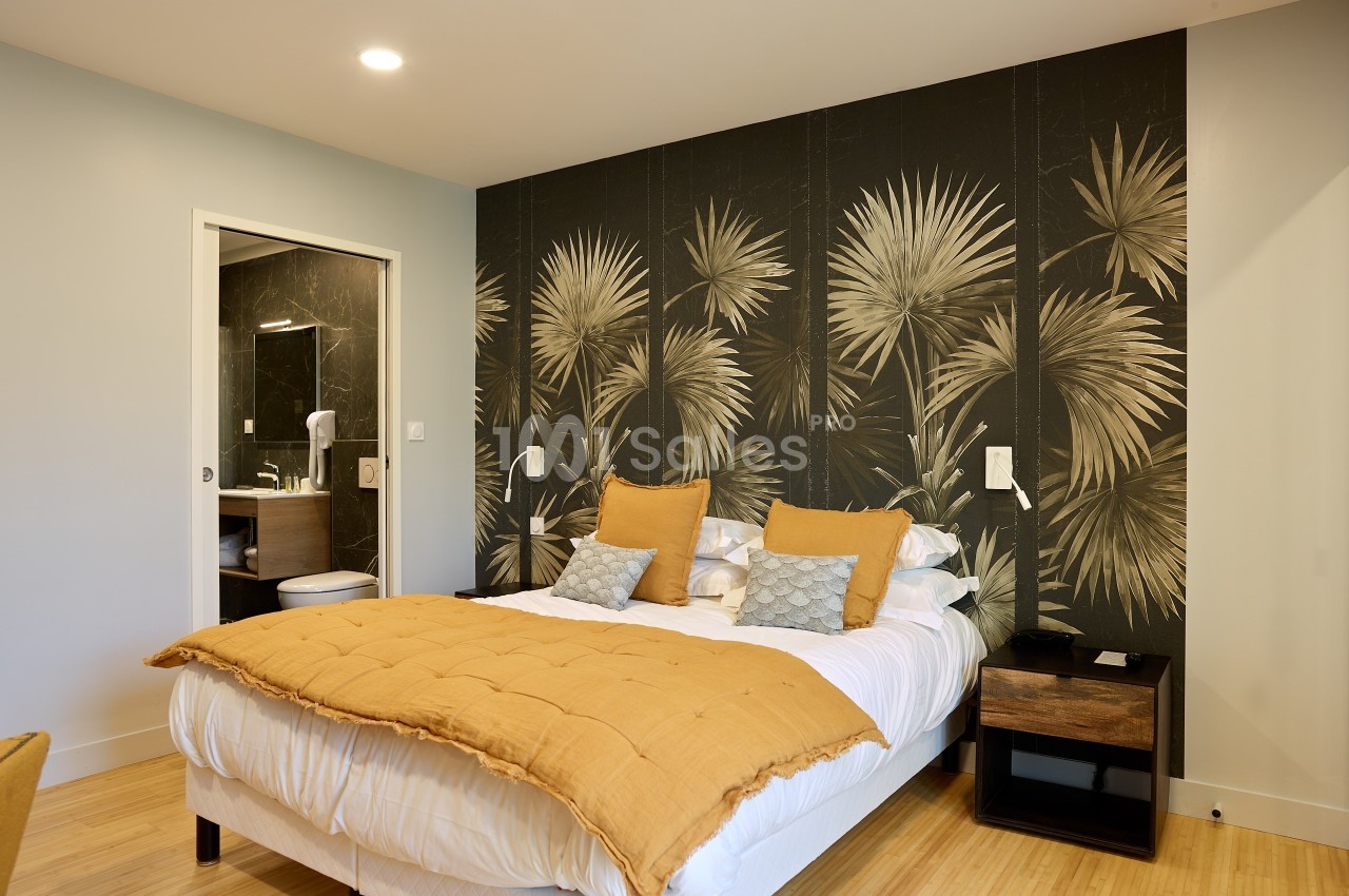 Chambre moderne avec lit double, couvre-lit jaune, mur décoré de motifs végétaux et salle de bain attenante visible.