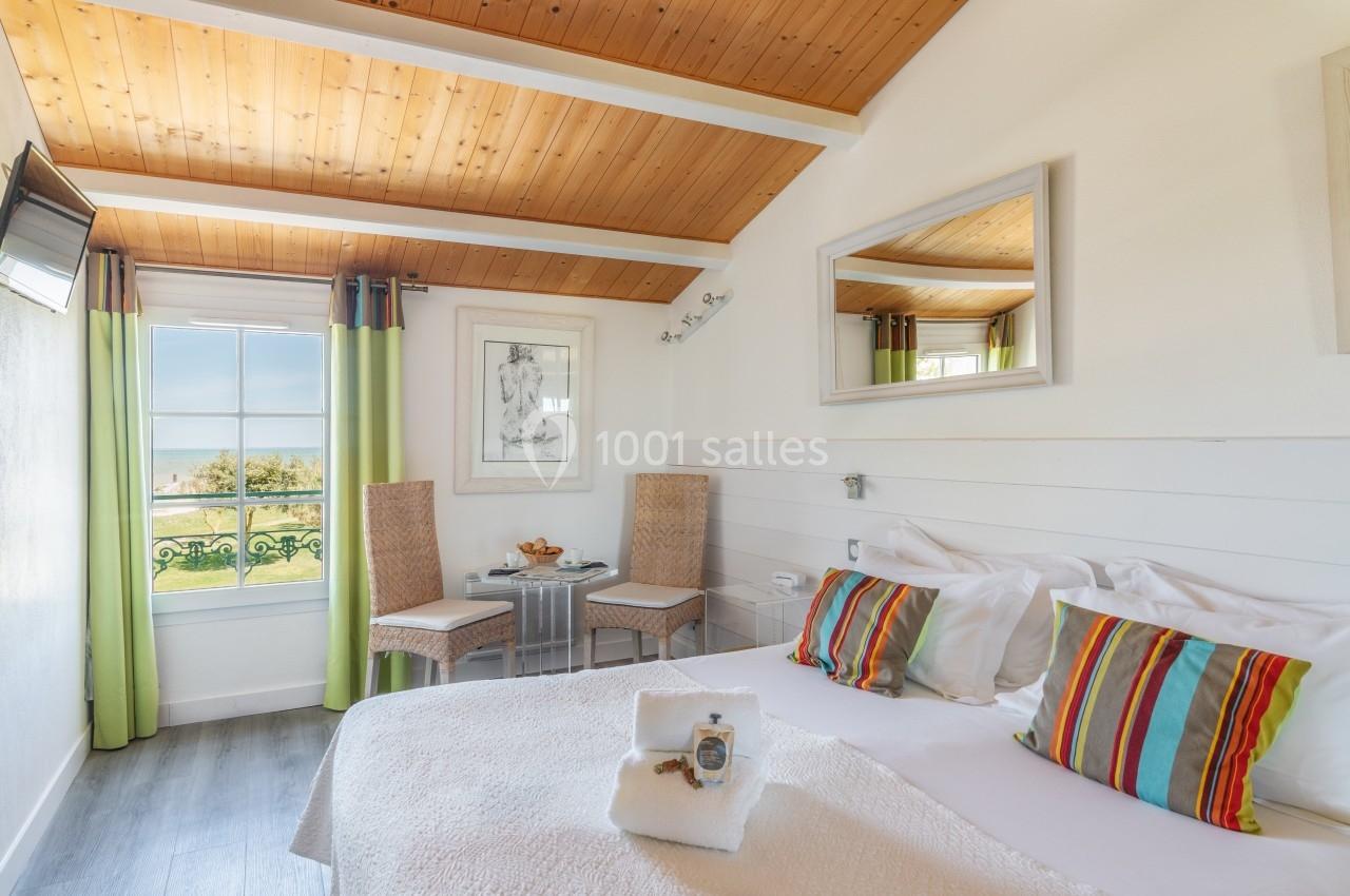 Chambre lumineuse avec lit double, plafond en bois, fenêtre donnant sur un paysage, et coin salon avec deux chaises.