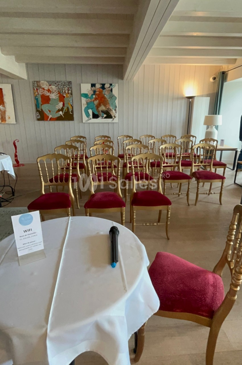 Salle de réunion avec des chaises dorées à assise rouge, une table ronde au premier plan et des tableaux accrochés au mur.