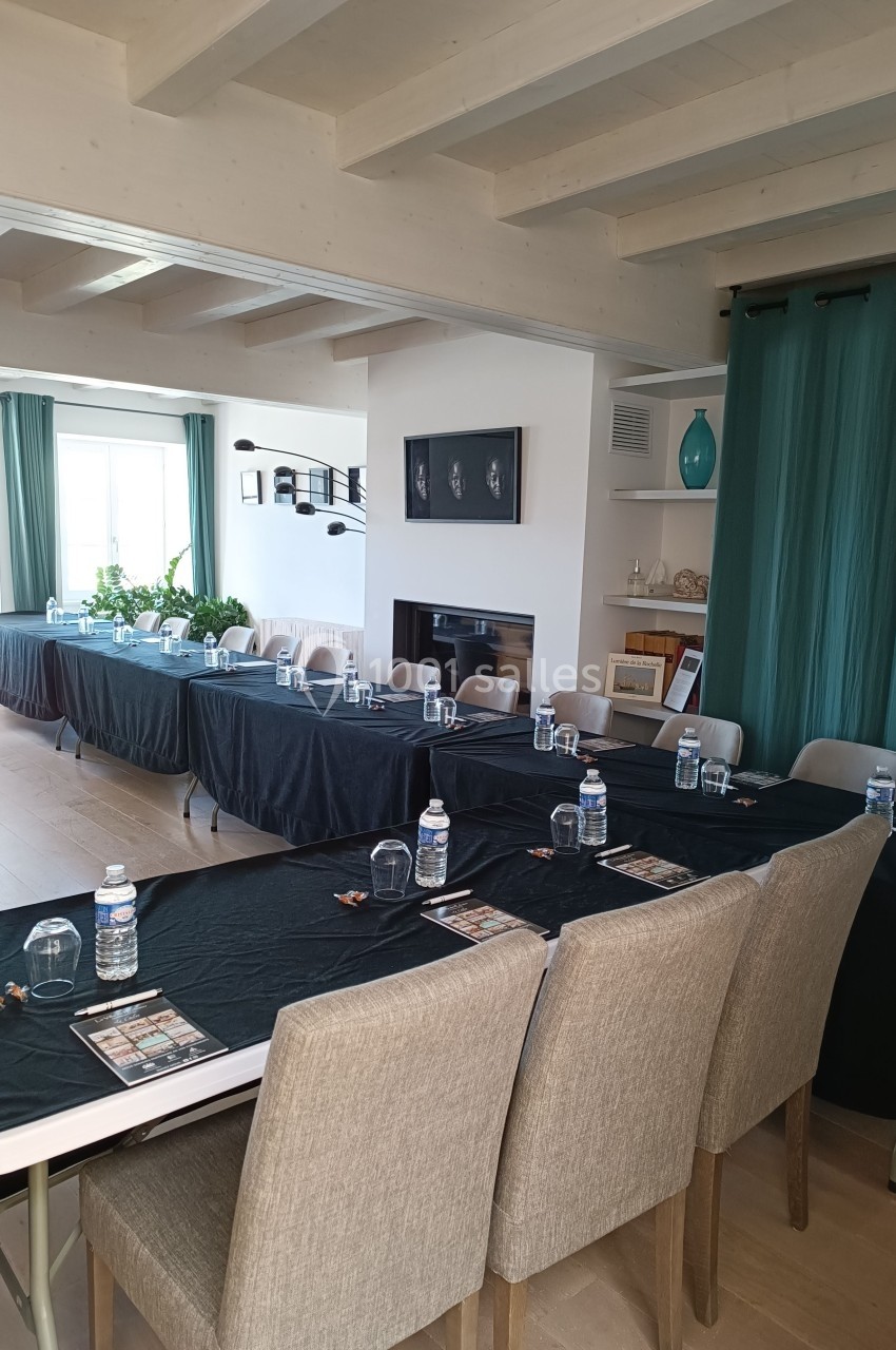 Salle lumineuse aménagée avec des tables noires alignées, des chaises grises et des bouteilles d'eau pour une réunion.