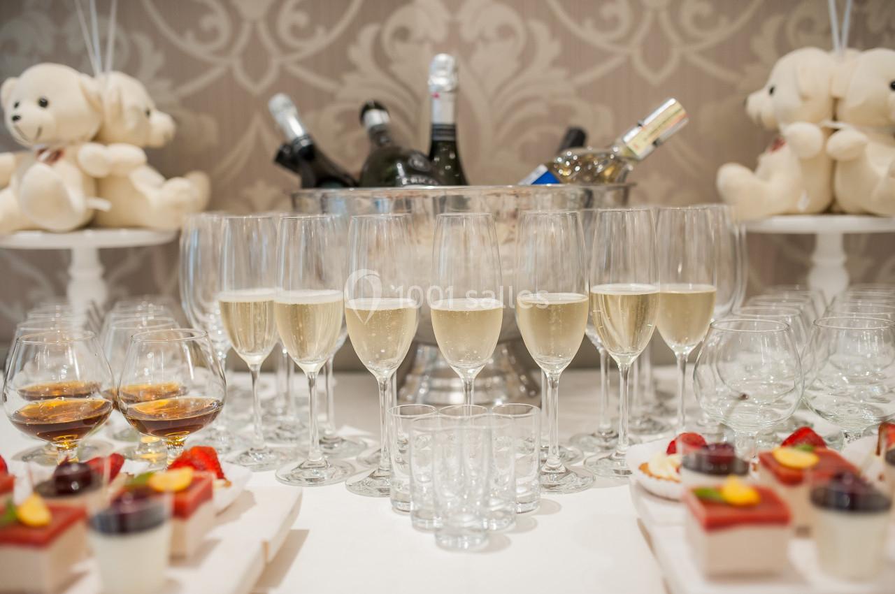 Verres de champagne alignés devant un seau à glace rempli de bouteilles, avec desserts et peluches en arrière-plan.