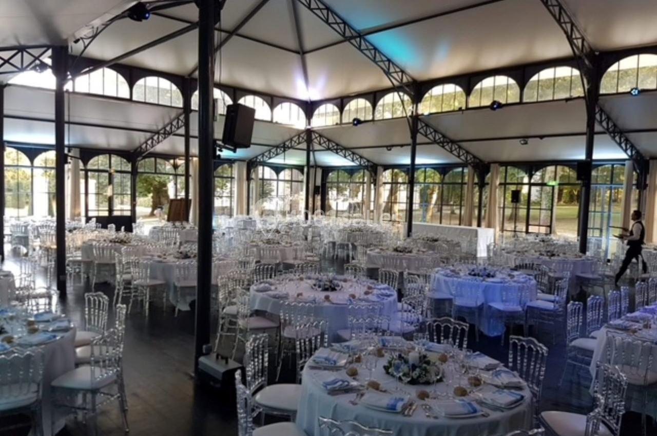 Salle de réception lumineuse avec des tables rondes dressées, chaises transparentes et structure métallique apparente.