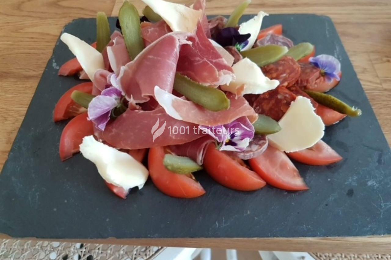 Assiette de charcuterie avec jambon cru, cornichons, fromage, tomates et fleurs comestibles sur une ardoise.