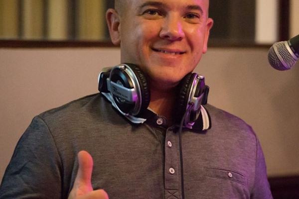 Homme souriant portant un casque audio autour du cou, levant le pouce devant un microphone.