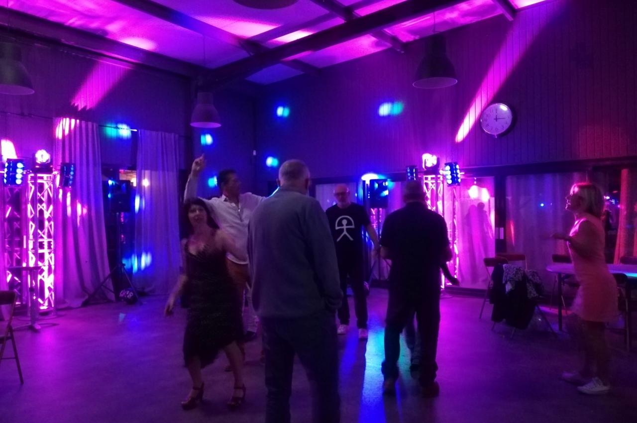 Personnes dansant dans une salle éclairée par des lumières colorées, avec des équipements de son et lumière en arrière-plan.