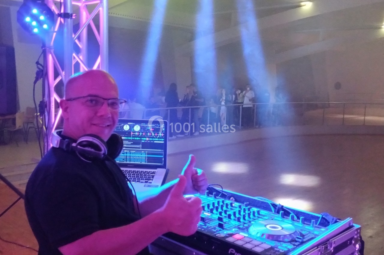 Un DJ souriant devant sa table de mixage, avec des lumières colorées et une piste de danse en arrière-plan.
