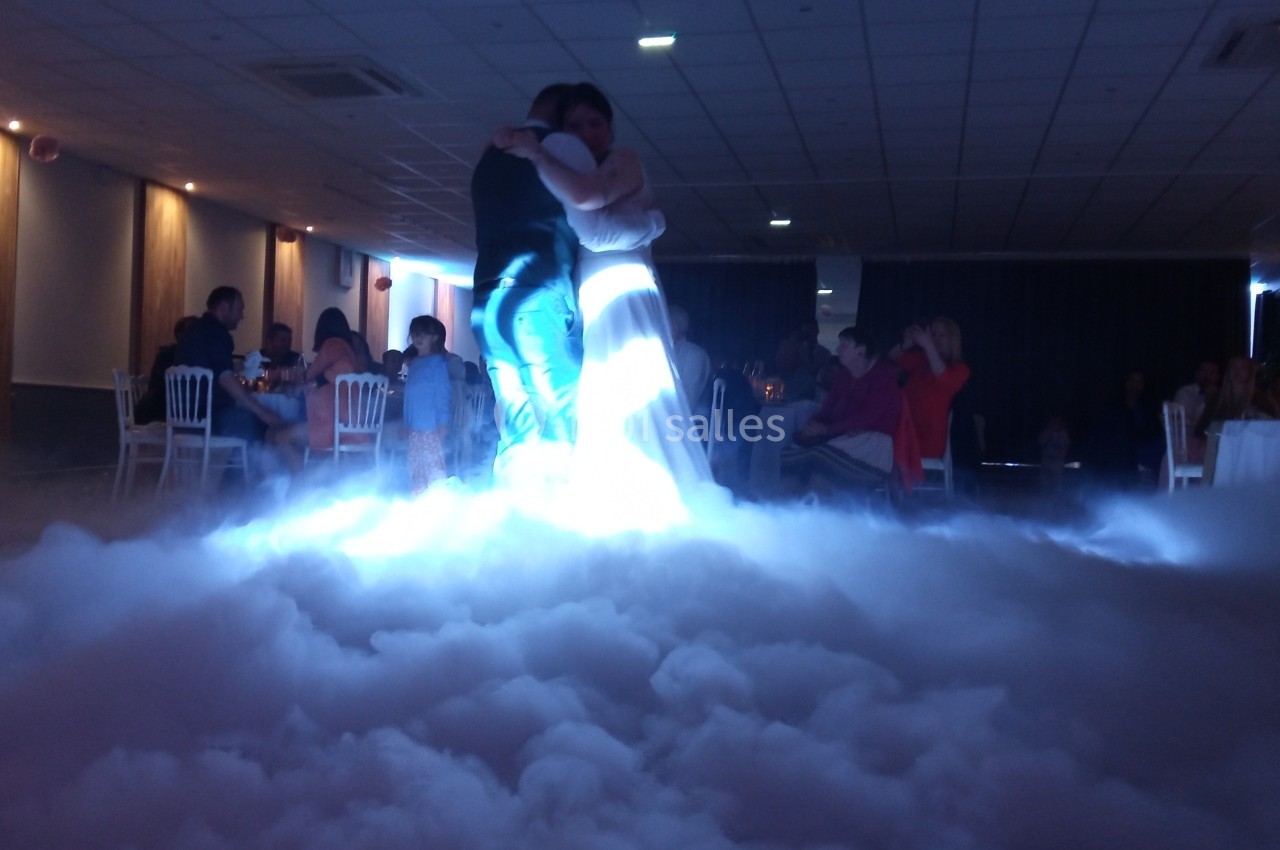 Un couple danse au centre d'une salle éclairée, entouré d'un effet de fumée au sol et de spectateurs assis.