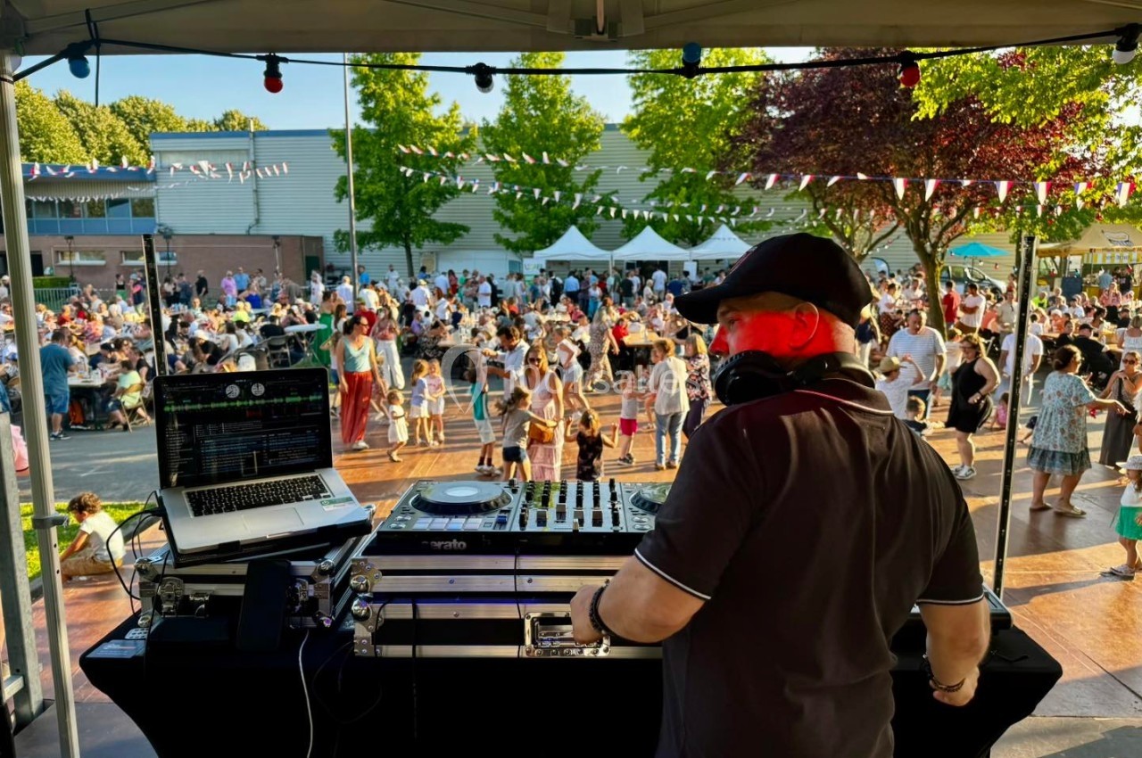 Un DJ mixe sur une scène en plein air, face à une foule rassemblée lors d'un événement festif en journée.