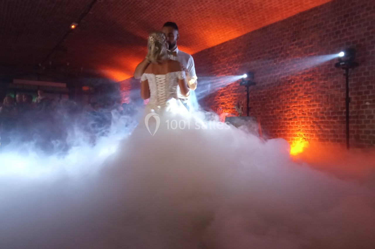 Un couple danse dans une salle en briques éclairée par des lumières chaudes, entouré d'un épais brouillard au sol.