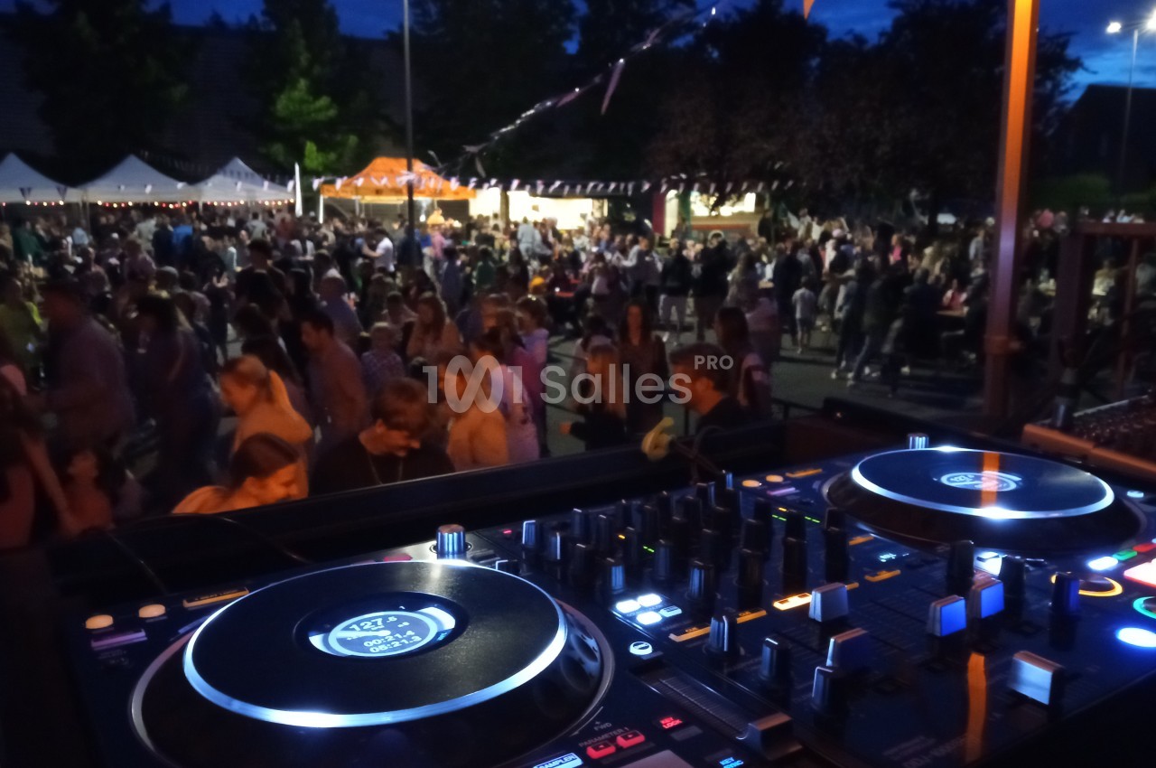 Vue d'une table de mixage en premier plan avec une foule rassemblée lors d'un événement nocturne en extérieur.