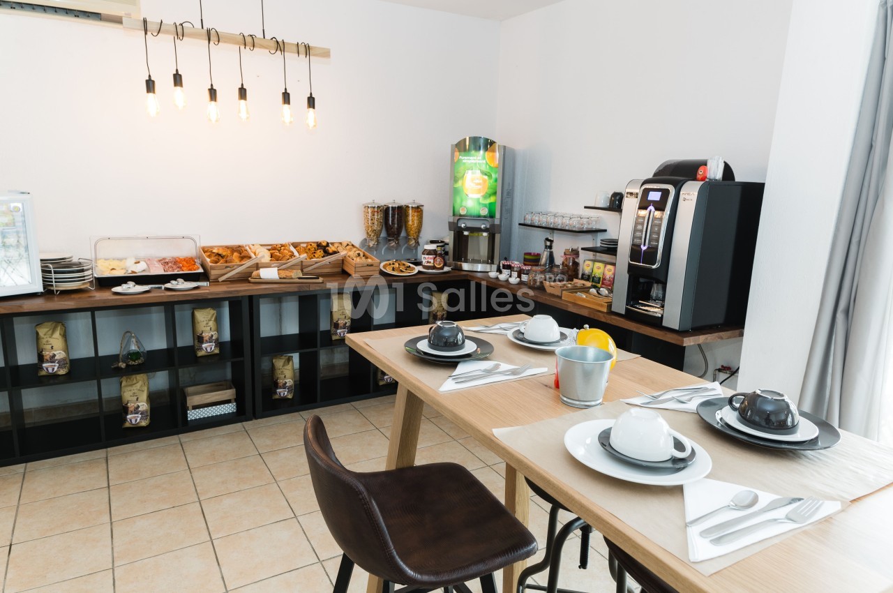 Salle de petit-déjeuner avec une table dressée de tasses et d'assiettes blanches, un buffet de viennoiseries et une machine à café à l'arrière-plan.