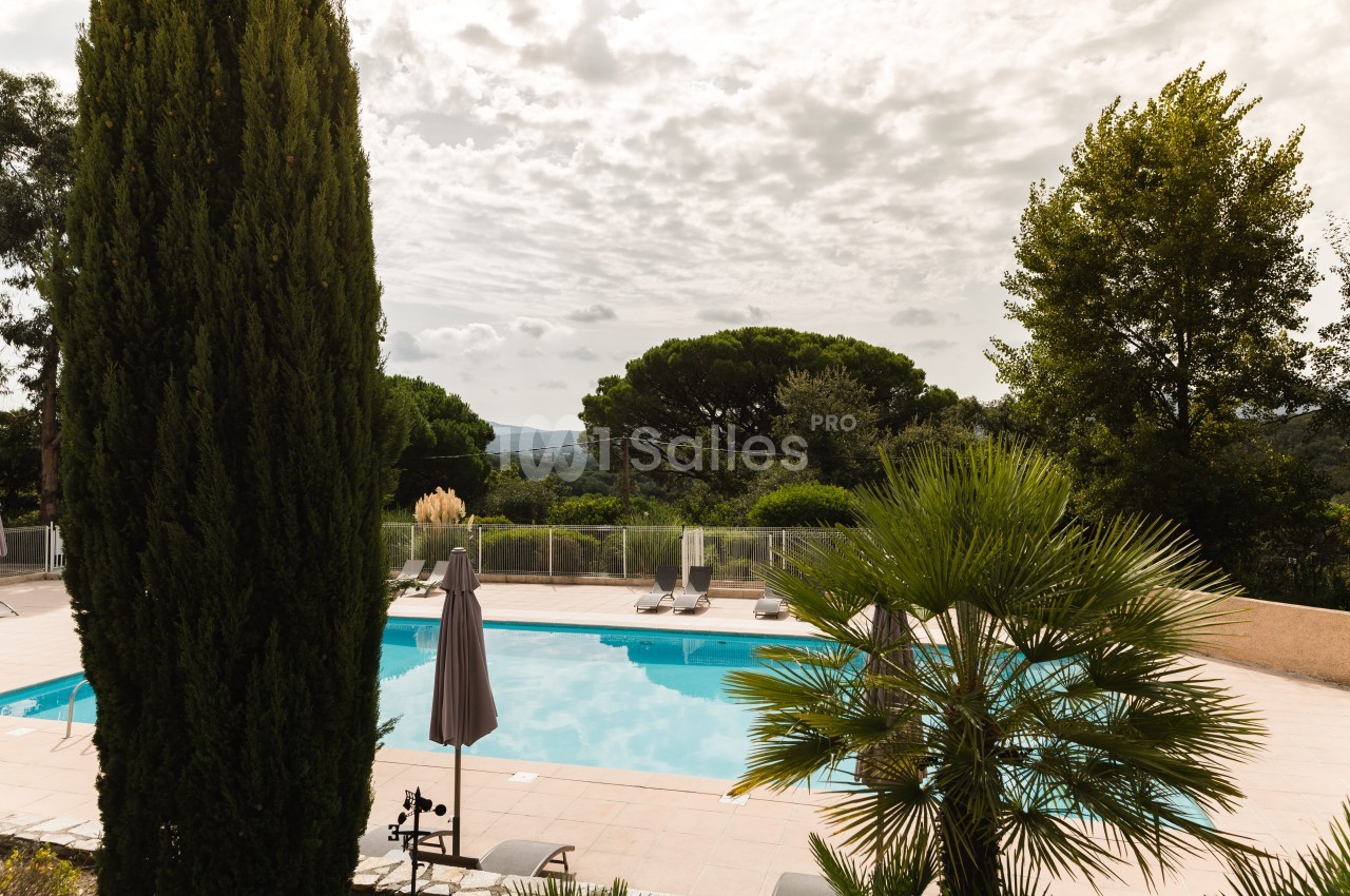 Piscine extérieure rectangulaire bordée de transats et d'un parasol fermé, entourée de cyprès, de pins et d'un palmier, sous un ciel nuageux.