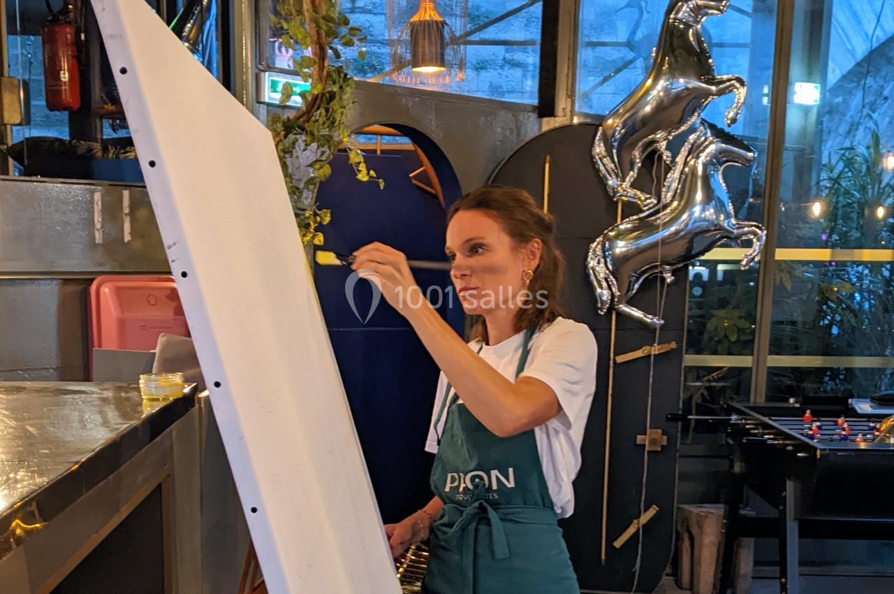 Une femme peint sur une grande toile blanche dans un espace intérieur décoré de plantes et de ballons argentés.