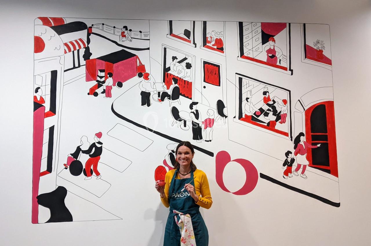 Fresque murale illustrant une scène urbaine animée avec des personnages, devant laquelle pose une personne souriante.