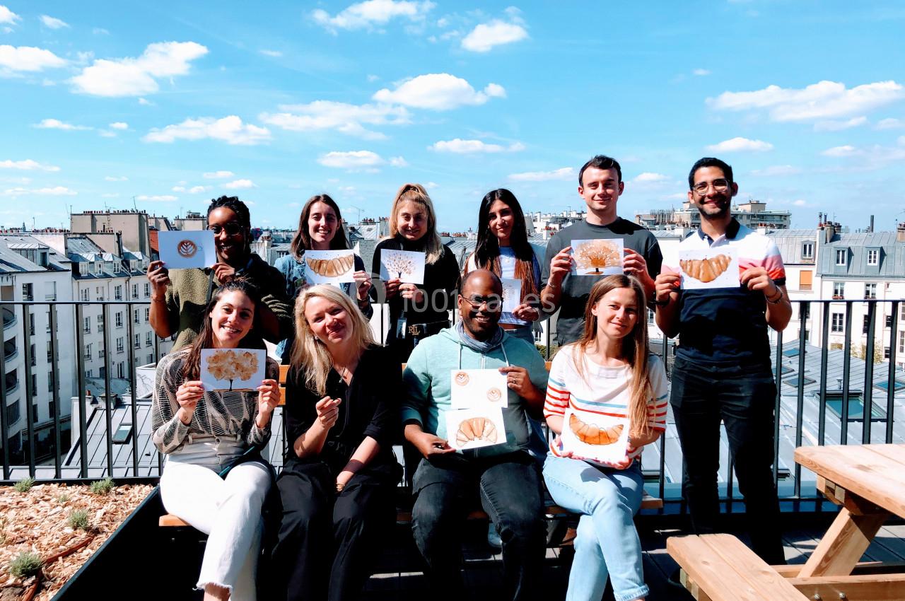 Un groupe de personnes souriantes tenant des dessins de croissants, assis sur une terrasse ensoleillée avec vue sur des…