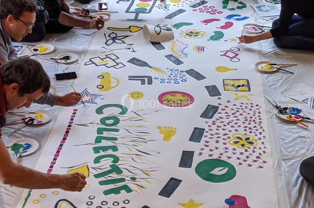 Des personnes peignent ensemble une grande fresque colorée avec des motifs variés et le mot ’COLLECTIF’ au centre.