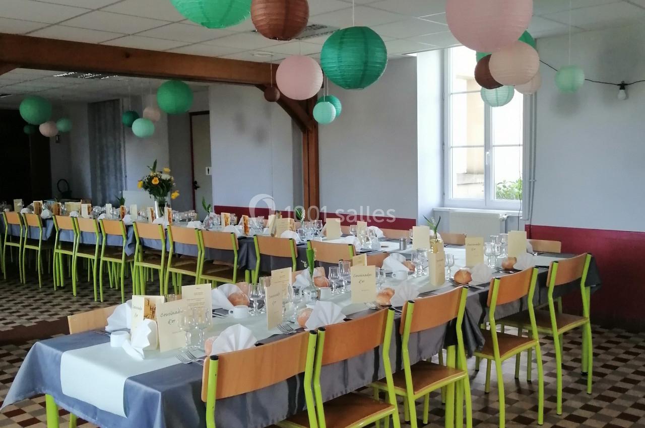 Salle décorée pour un événement, avec tables alignées, nappes grises, chaises vertes et lanternes suspendues colorées.