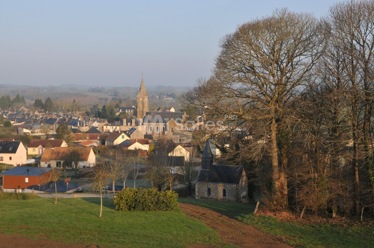 Village rural avec église au centre, maisons environnantes, arbres dénudés et paysage vallonné en arrière-plan.