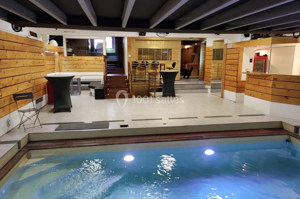 Piscine intérieure avec éclairage, entourée de murs en bois et d'un espace aménagé avec tables et chaises.