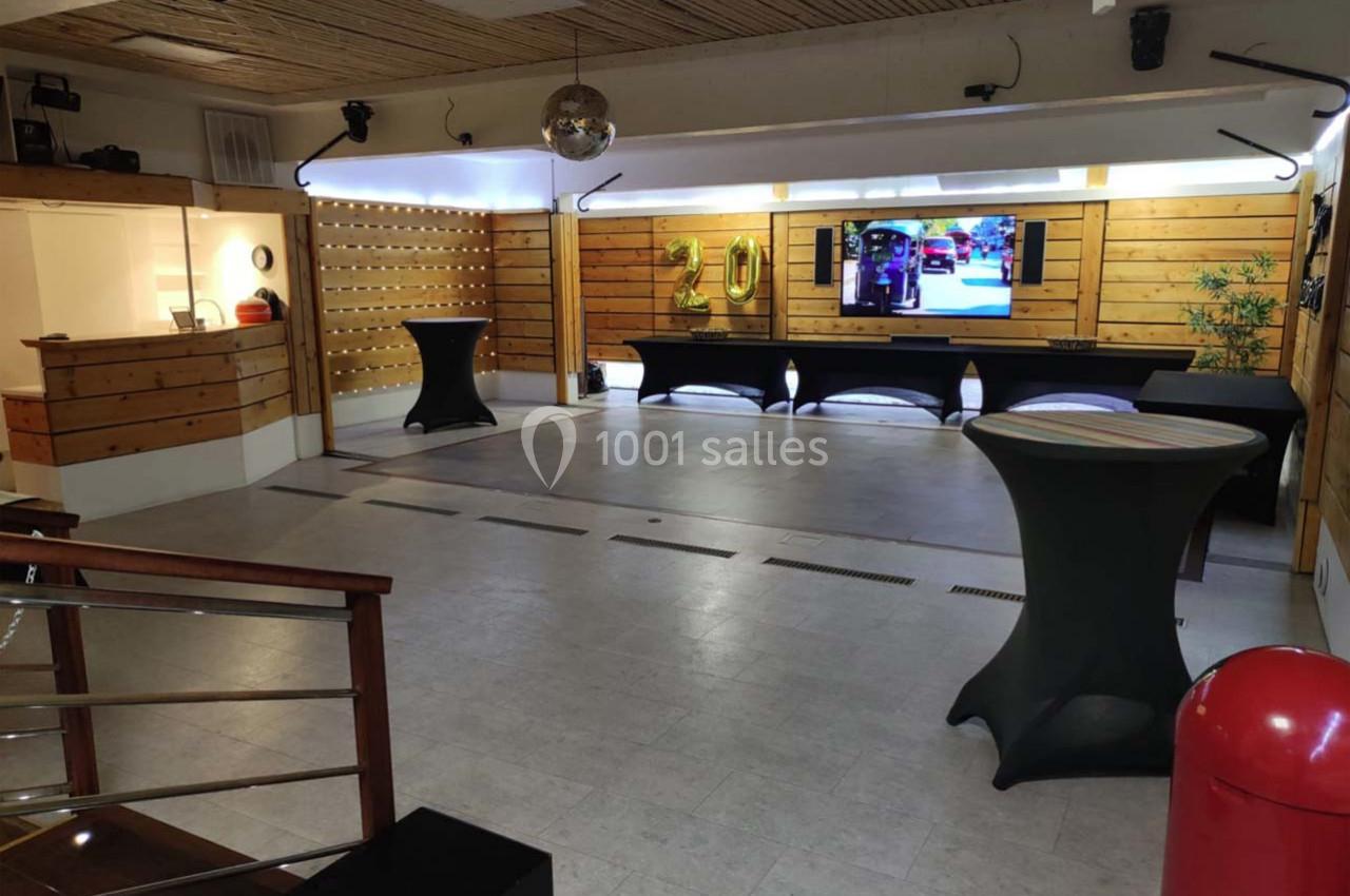 Salle vide avec parquet au plafond, tables hautes habillées de housses noires, écran mural et décoration en bois.