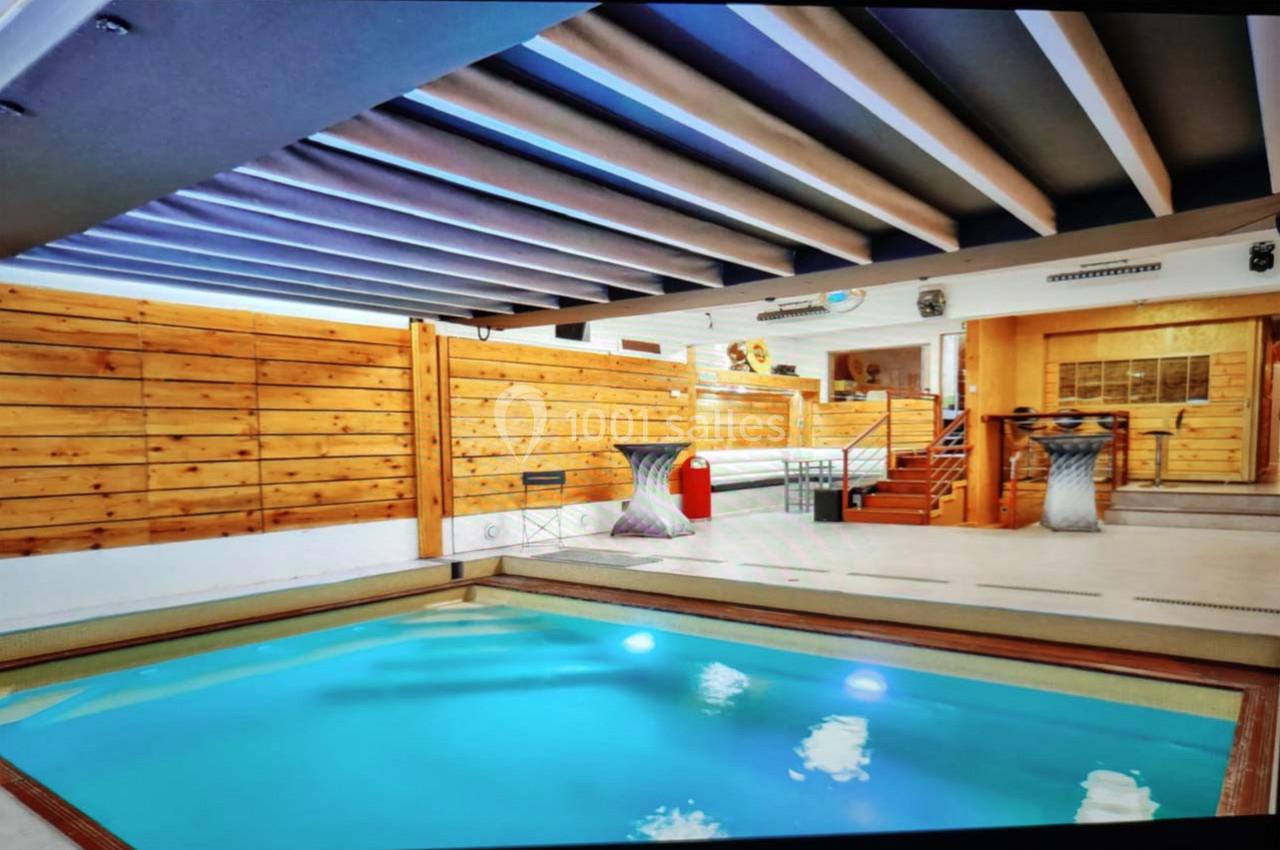 Piscine intérieure avec murs en bois, plafond à poutres apparentes et espace aménagé avec escaliers et mobilier.